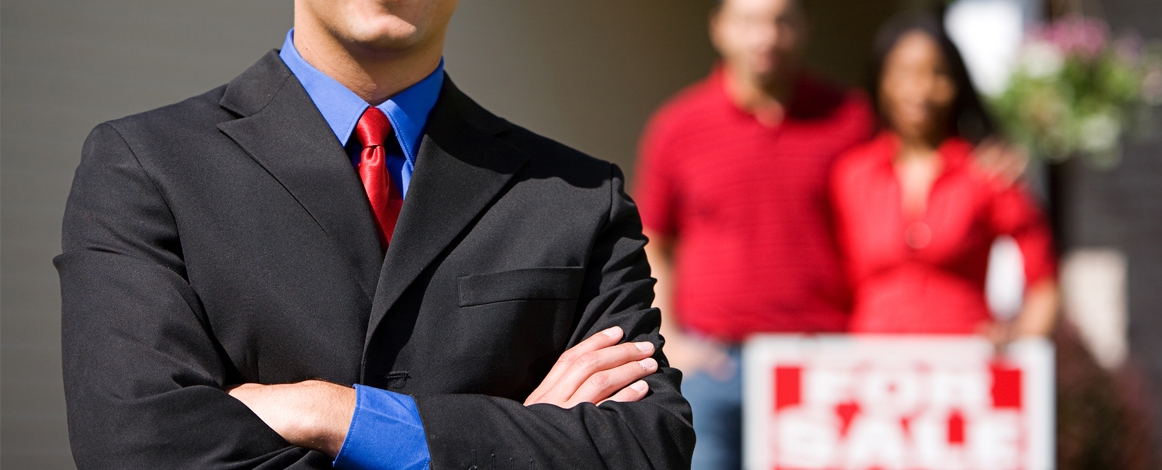 White male REALTOR in the foreground with sellers in background in front of for sale sign